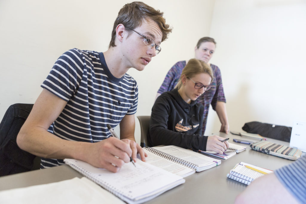 A male student takes notes during a class while two other individuals participate in the background.