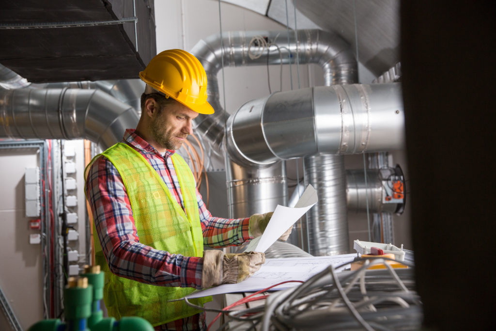 Man in yellow safety hat and safety vest reading blueprints in a room with HVAC equipment.
