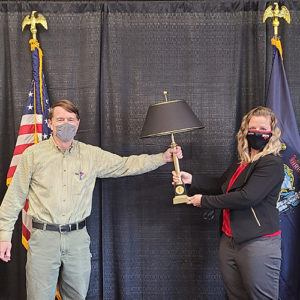 Don Varney and Dr. Libby pose wtih a black lamp on a black cloth backdrop. The State of Maine flag and the United States flag are behind them.