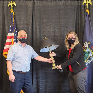 Ray Masse and Dr. Libby pose wtih a black lamp on a black cloth backdrop. The State of Maine flag and the United States flag are behind them.