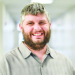 Man with salt and pepper hair and a brown beard, wearing a light gray plaid button down shirt.