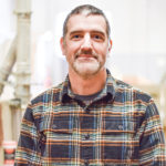 Man with salt and pepper hair and beard, wearing a navy, light blue, tan and white plaid flannel over a gray shirt.