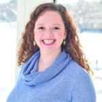 Woman with shoulder length auburn curly hair and brown eyes, wearing a light blue cowl-neck sweater.