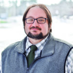 Man with brown hair and a beard and black glasses, wearing a gray vest over a plaid shirt and green tie.