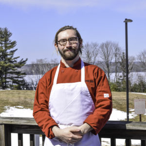 Benjamin Simpson stands outside in his red chef's coat and white jacket. Benjamin has brown hair, a beard and mustache, and glasses.