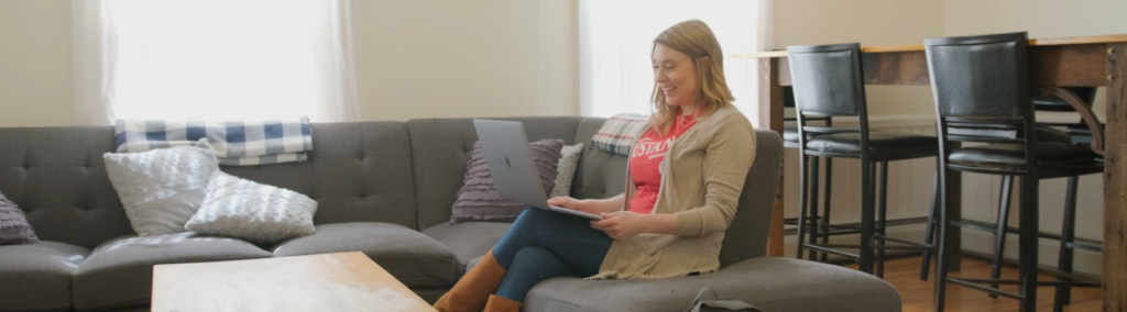 A female student is sitting on a gray sectional sofa with a laptop in her lap, looking at the screen.