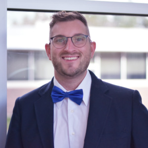 Man with cropped haircut and glasses, wearing a navy blue jacket over a white shirt with a royal blue bow tie.